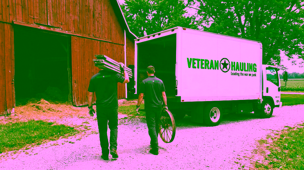 Two crew members in dark polos, seen from behind, walk across a gravel lane from an open red barn toward a white Veteran Hauling box truck with its back door open, one carrying weathered barn boards on his shoulder and the other an old rusted iron wheel.