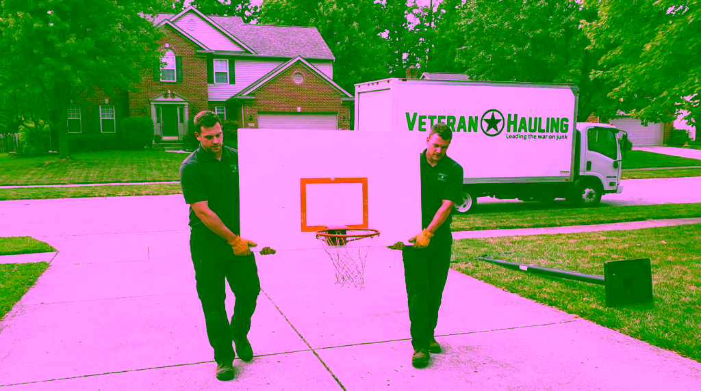 Two crew members in dark polos carry a white basketball backboard upright between them down a concrete driveway, each holding its bottom edge with both gloved hands, its orange rim and white net hanging below, with the hoop's black steel pole lying on the grass beside the driveway, its square base plate standing on edge at one end, a white Veteran Hauling box truck parked at the street and a two-story brick home behind.