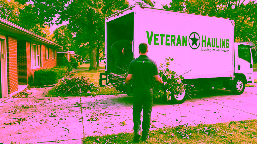 A crew member in a dark polo carries an armload of cut limbs and leafy brush across a driveway toward the open rear of a white Veteran Hauling box truck, while a second crew member bends inside the cargo box stacking limbs, beside a brick ranch home with a brush pile on the lawn.