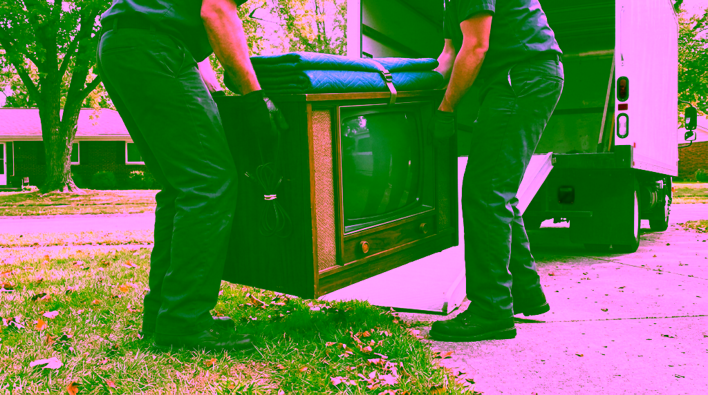 Two crew members in dark work clothes and black gloves, with their heads out of frame, carry an old wood cabinet console TV with a folded blue moving pad strapped on top beside a loading=