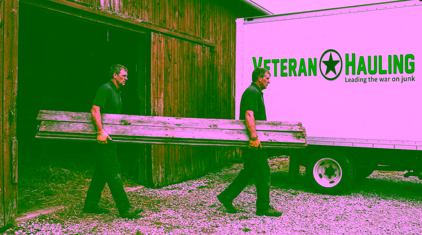 Two crew members in dark polos carry a long stack of weathered gray barn boards out of an open red barn onto gravel, one at each end, toward a white Veteran Hauling box truck, with loose hay on the barn floor behind them.