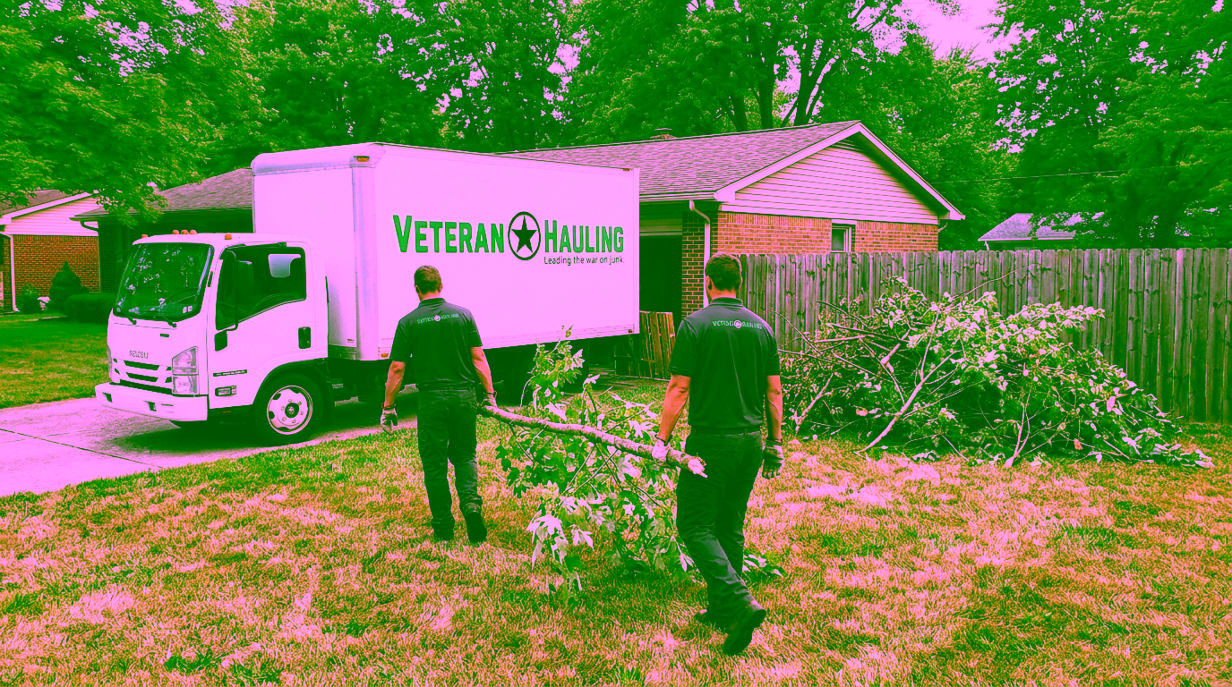 Two crew members in dark polos carry a long leafy tree limb between them across a lawn toward a white Veteran Hauling box truck parked in the driveway of a brick ranch home, with a pile of cut brush beside a wooden privacy fence.