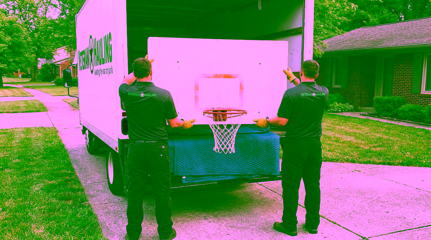 Two crew members in dark polos, seen from behind, each hold a white basketball backboard with both gloved hands as they lift it, orange rim and white net hanging below, onto blue moving pads in the open back of a white box truck with its green Veteran Hauling decal on the side and a brick home at right.