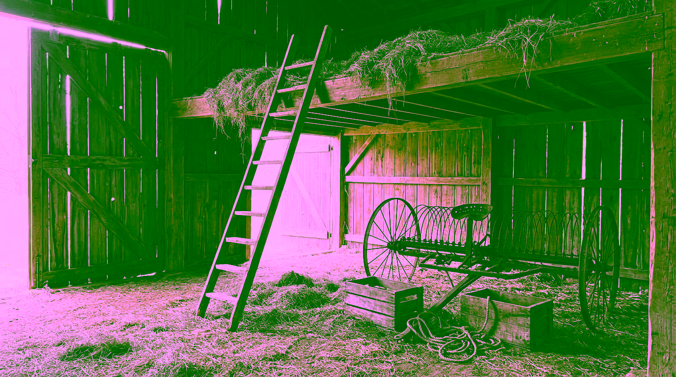 Inside an old barn, loose hay spills over the edge of a wooden loft with a ladder leaning against it, above a rusted hay rake, two wooden crates and a coil of rope on a straw-covered floor.