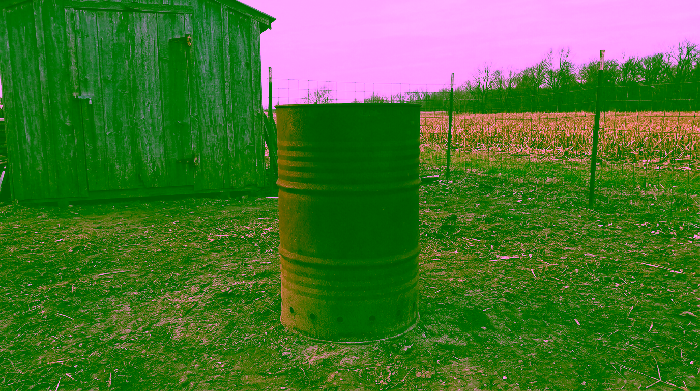 A rusty steel barrel with vent holes near its base stands on bare dirt beside a weathered wooden shed, with a wire fence and a harvested cornfield behind it.